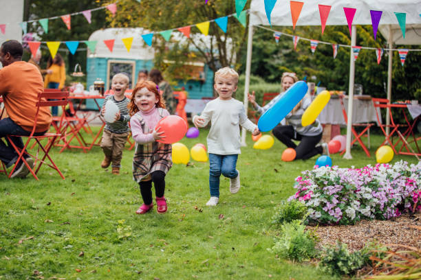 Three young children enjoy playing with balloons at a garden party as they run towards the camera. Adults are sat talking in the background and looking at the children. There is a food truck in the background and a marquee and colorful bunting fill the garden